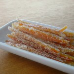 Photo of straight strips of candied orange peels rolled in sugar in a white dish resting on a table.